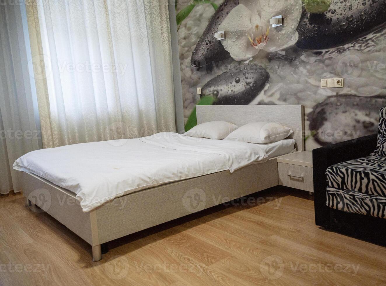 double bed with two pillows, next to it is a chair. double bed with a bedside table. white bed linen. orchids on the photo wallpaper. next to the bed is a window and a chair