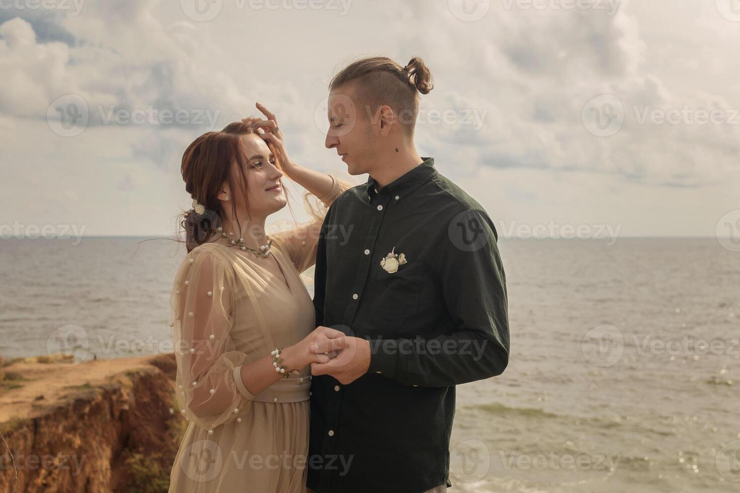 Beautiful lovers stand against the sky and sea and look at each other. Close-up against the sky with clouds. The girl has her hair beautifully styled in a bun, she adjusts her hair with one hand photo
