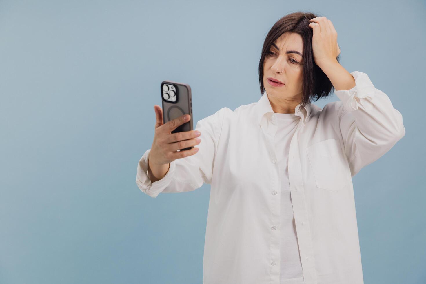 Confused mature woman scratching head while using smartphone on blue background photo