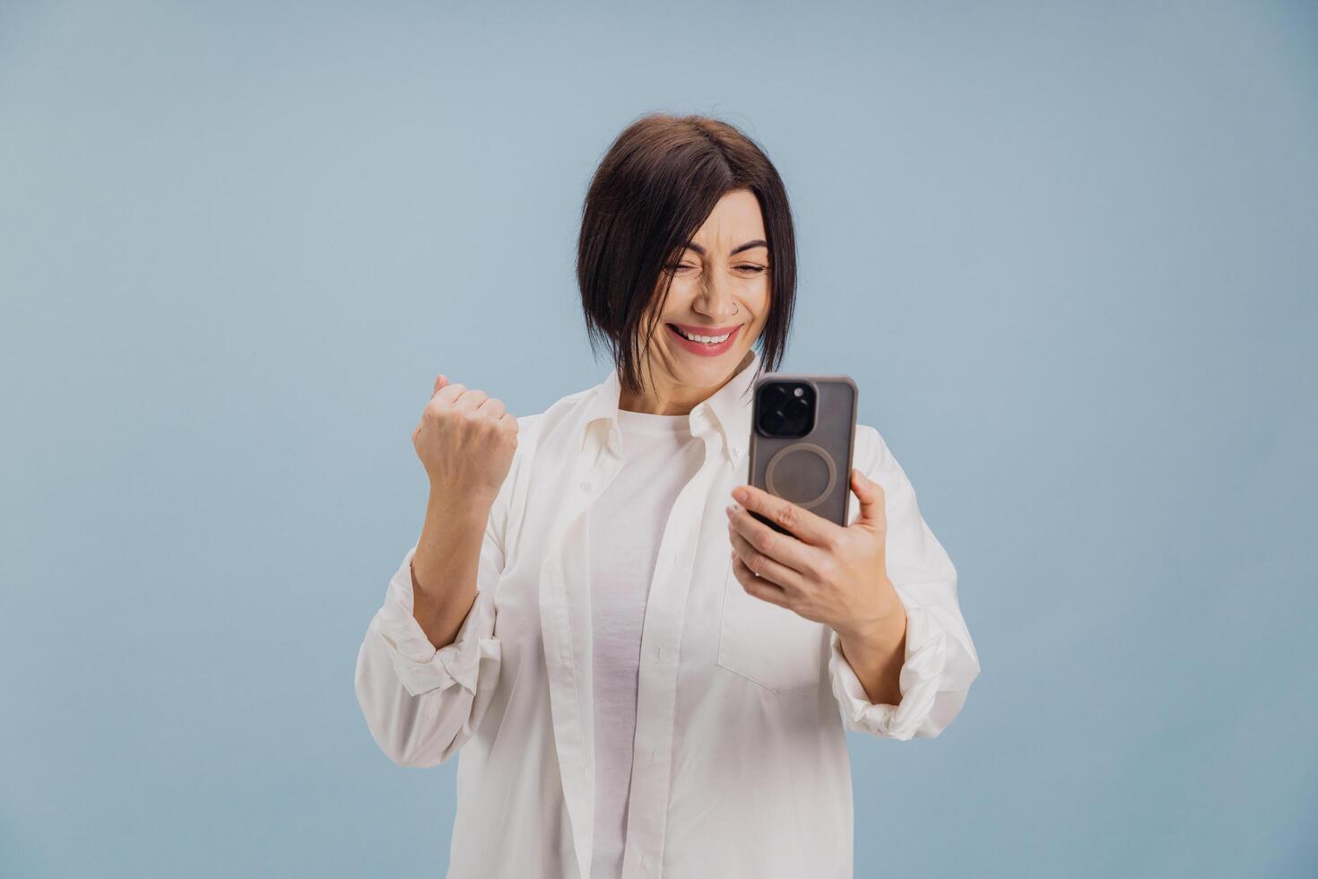 Excited mature woman celebrating success while using smartphone on blue background photo