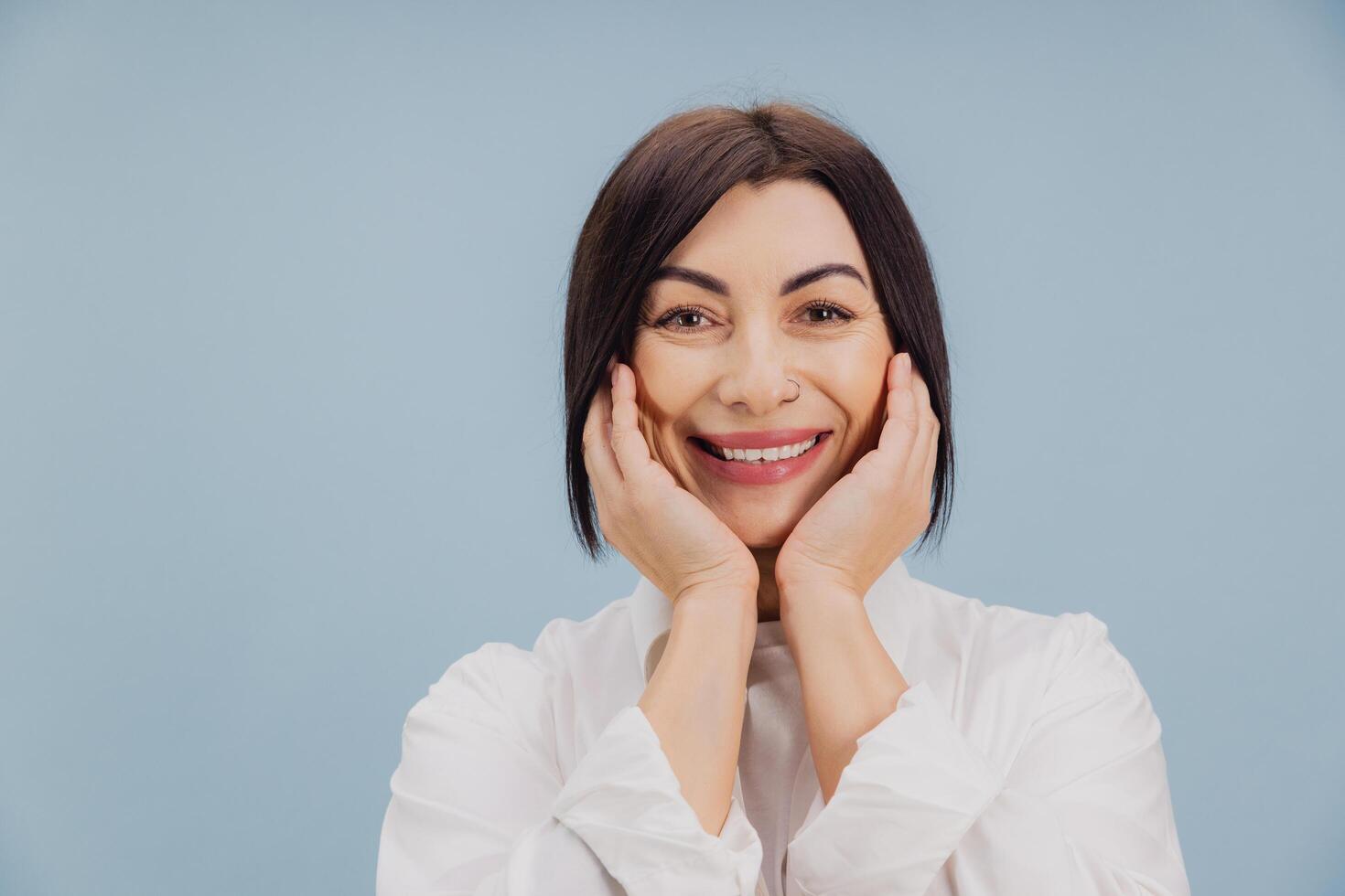Happy mature woman holding face with hands and smiling on blue background photo