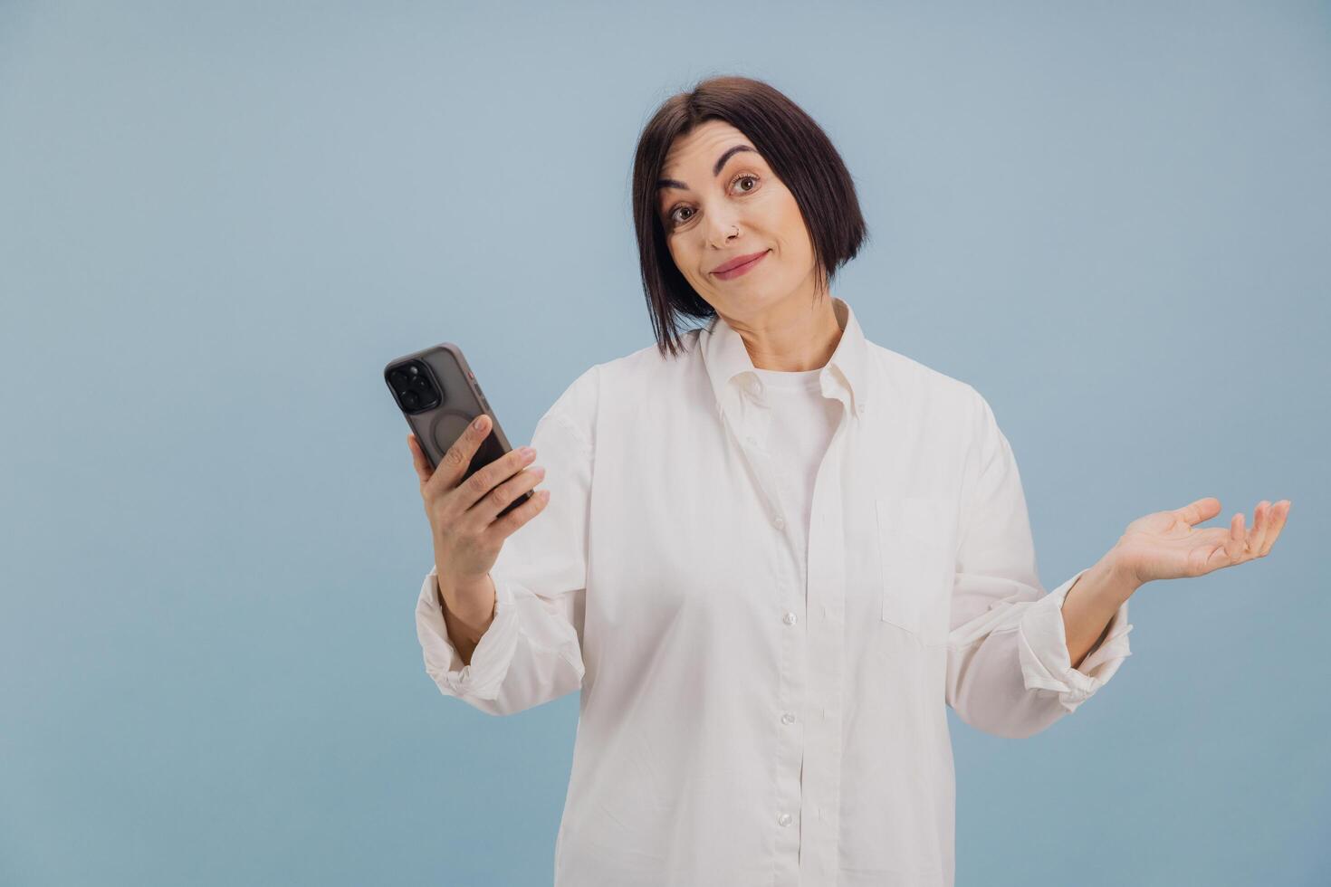 Brunette mature businesswoman holding smartphone shrugging shoulders on blue background photo