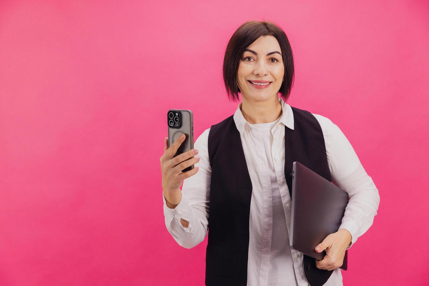Smiling businesswoman holding laptop and using smartphone on pink background photo