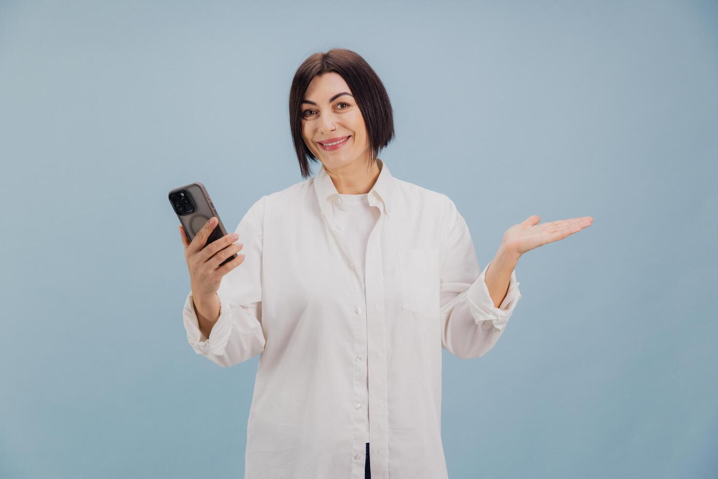 Smiling mature woman holding smartphone and showing something on light blue background photo