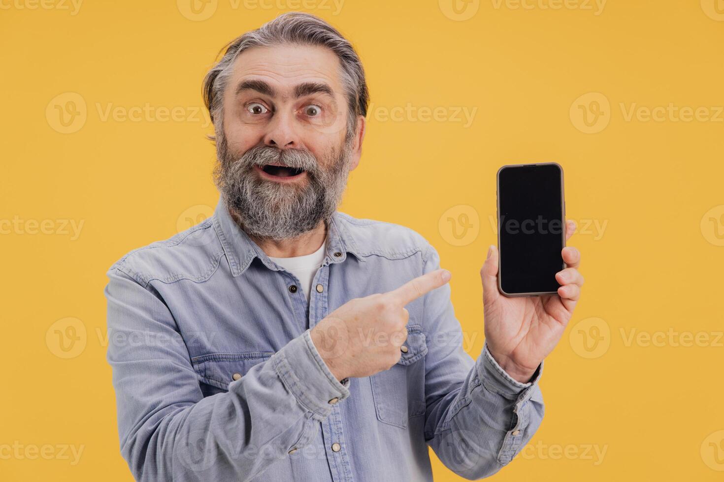 Surprised senior man pointing at smartphone with blank screen on yellow background photo
