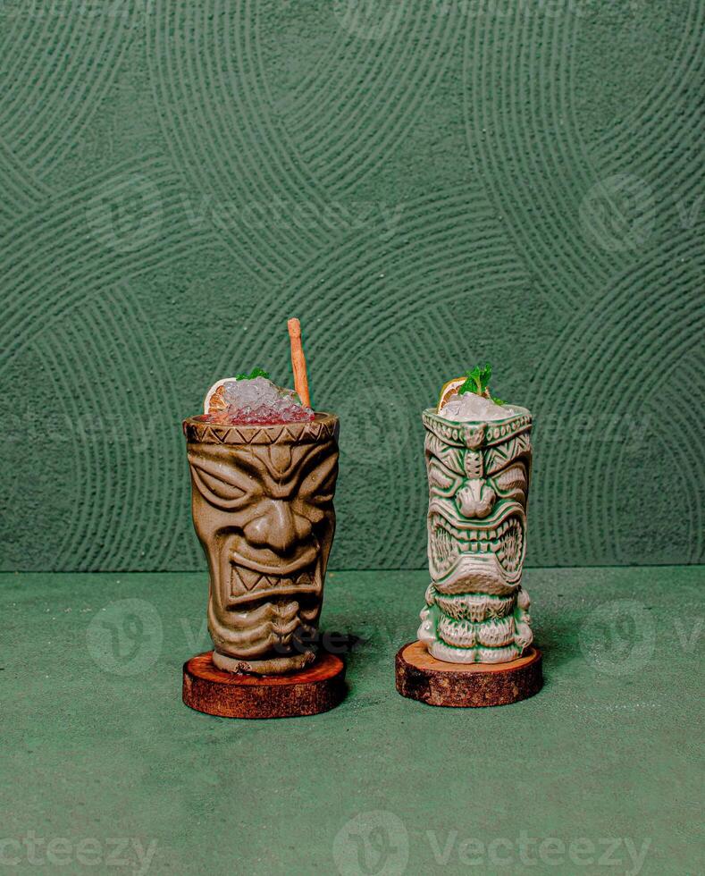 Refreshing Drinks In Two Tiki God Glasses, Elegantly Served On Round Wooden Coasters, Offering A Stylish And Tropical Vibe. photo