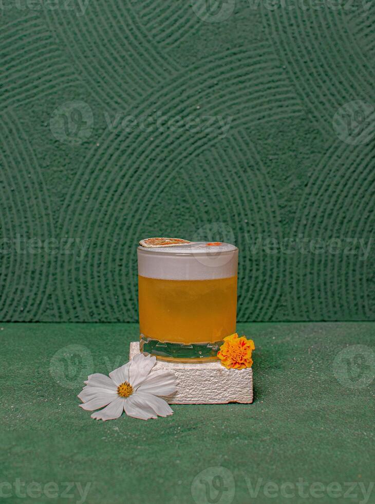 A Cocktail Glass Featuring A Round Ice Ball, Topped With A Decorative Garnish, Elegantly Served On A Minimalist White Block With Flowers. photo