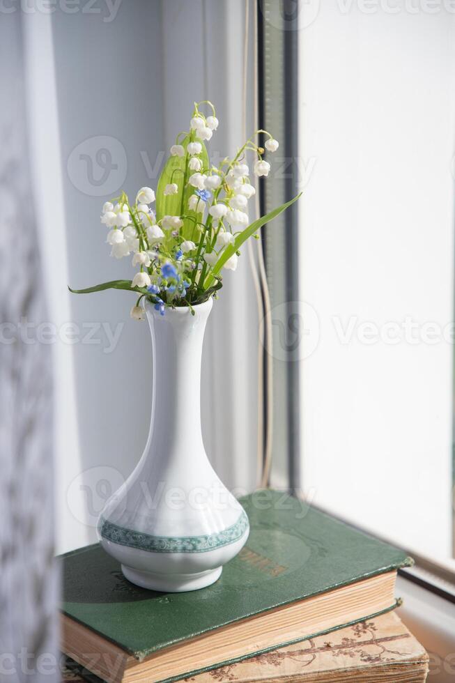 elegant small bouquet of lilies of the valley in a chinese porcelain vase in summer on a stack of old books on a windowsill, photo