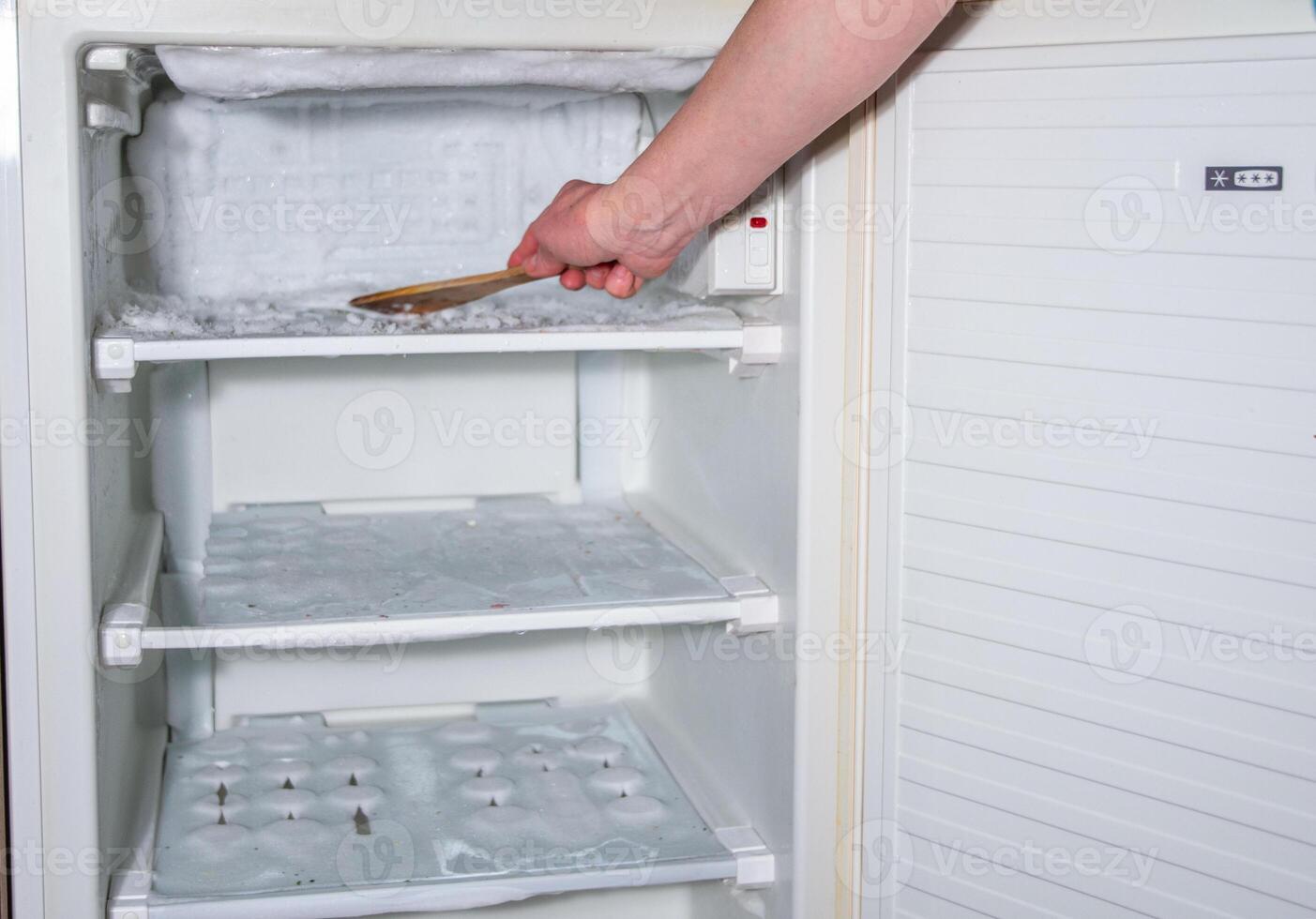 ice has formed on the walls of the freezer and it is creeping out, a woman's hand is knocking the ice off with a wooden spatula, photo
