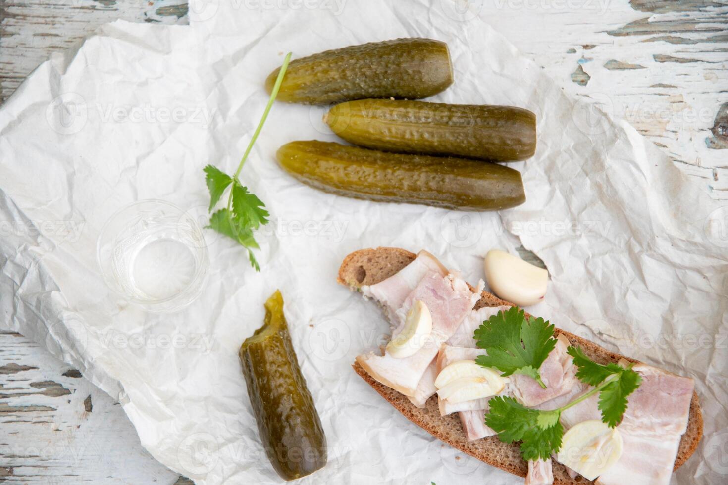 Rustic appetizer of pickles, rye bread with bacon, garlic and parsley on white paper, laid out on a weathered wooden surface with a shot of vodka, photo