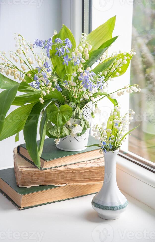 two bouquets of spring flowers on the windowsill, one of which stands on a stack of old books, lilies of the valley, creeping blue phlox, photo