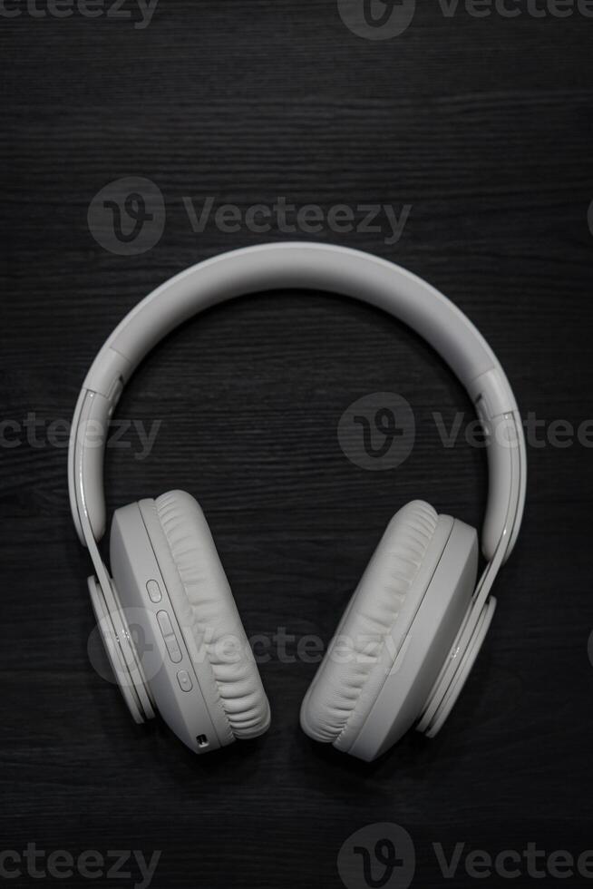 A pair of white over-ear wireless headphones with cushioned ear pads and control buttons on the left ear cup. Dark wooden background photo