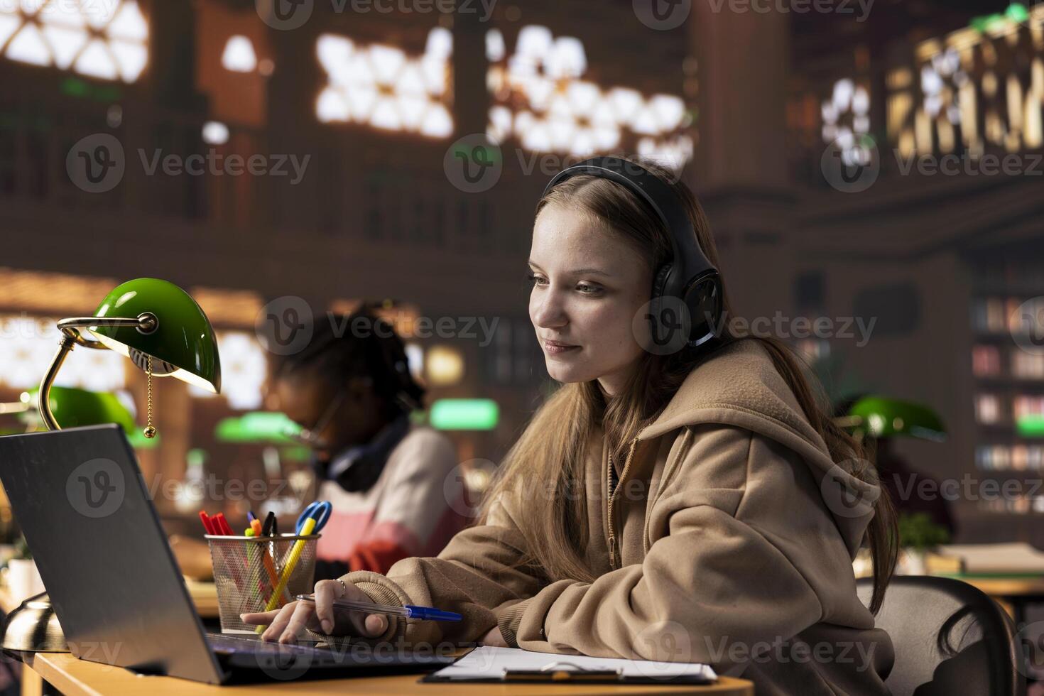 College student paying attention during remote lesson live event via call meeting, takes notes from the webinar discussion in the university library on campus. Pupil attends online course. photo