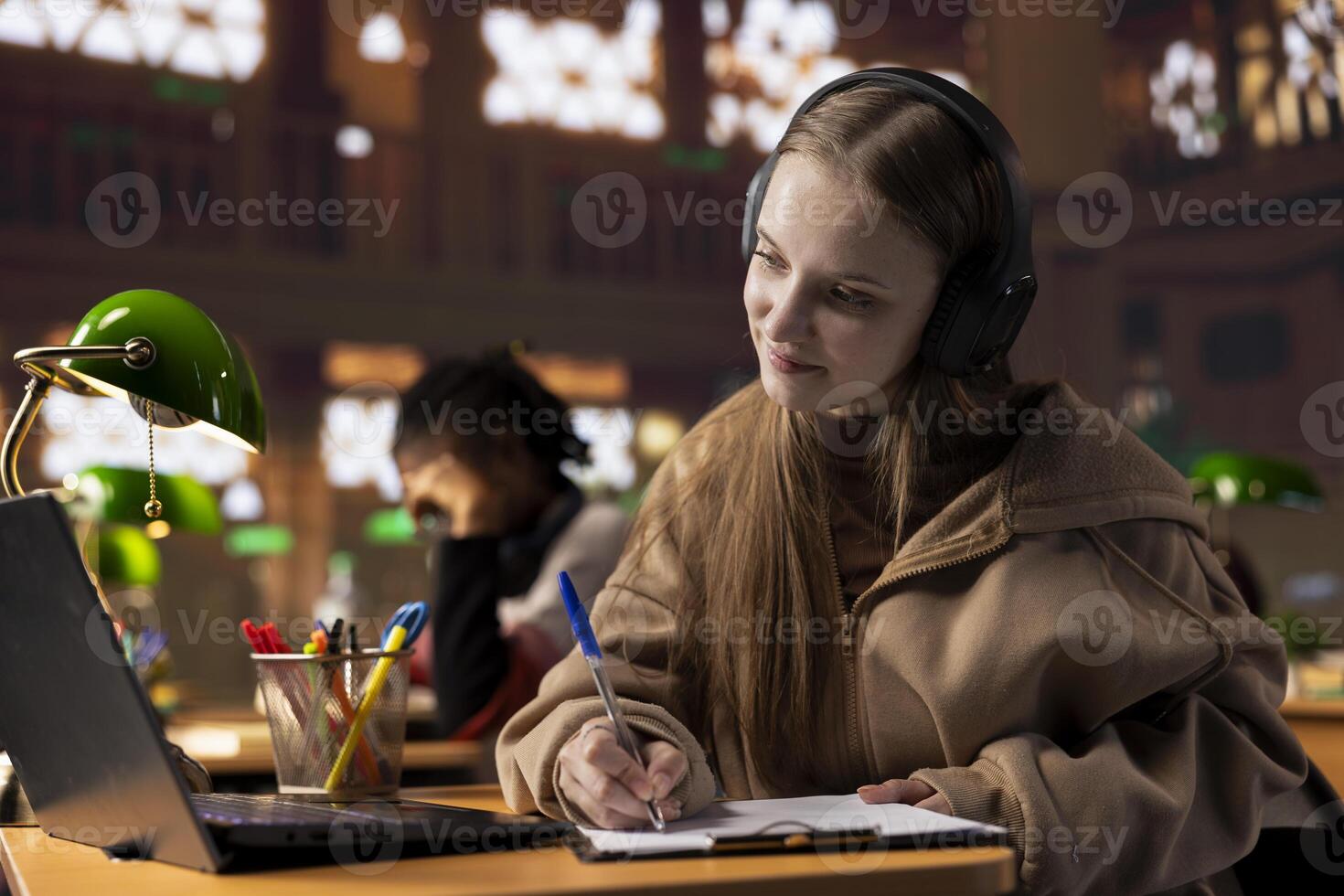 Young pupil takes notes from the teacher via conferencing webinar, attending online class from the campus library. University student writes information from the academic web lecture. photo