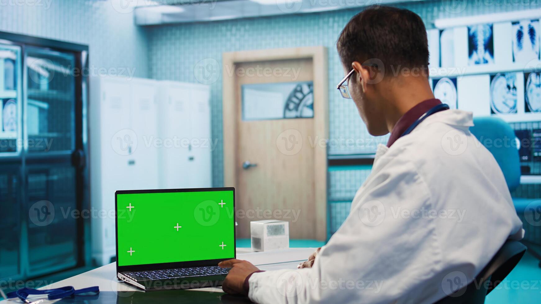 Indian physician works in his facility office next to chroma key, using modern technology for scan interpretation and results. Specialist offers professional advice based on records. Camera B. photo