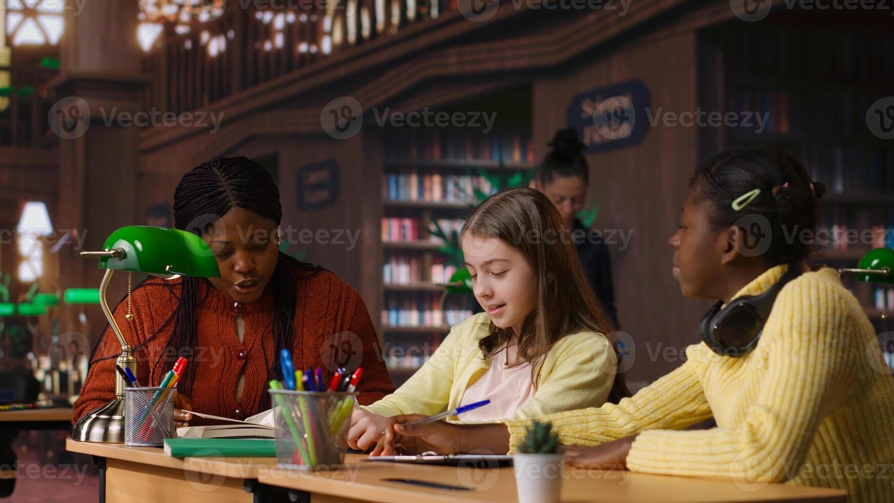 African american private teacher offering support to her students, tutoring them through a lesson plan at a library desk. Tutor providing learning materials, mentoring scholars. Camera B. photo