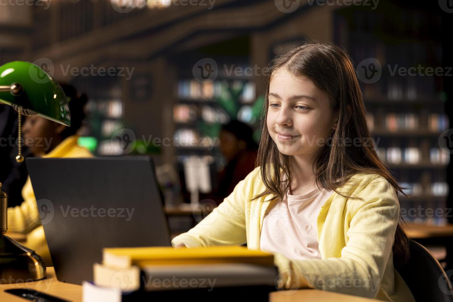 Primary school student sitting at desk in the campus library and writing on laptop, preparing notes and ideas for future lessons. Scholar girl using digital gadget for assignments. photo