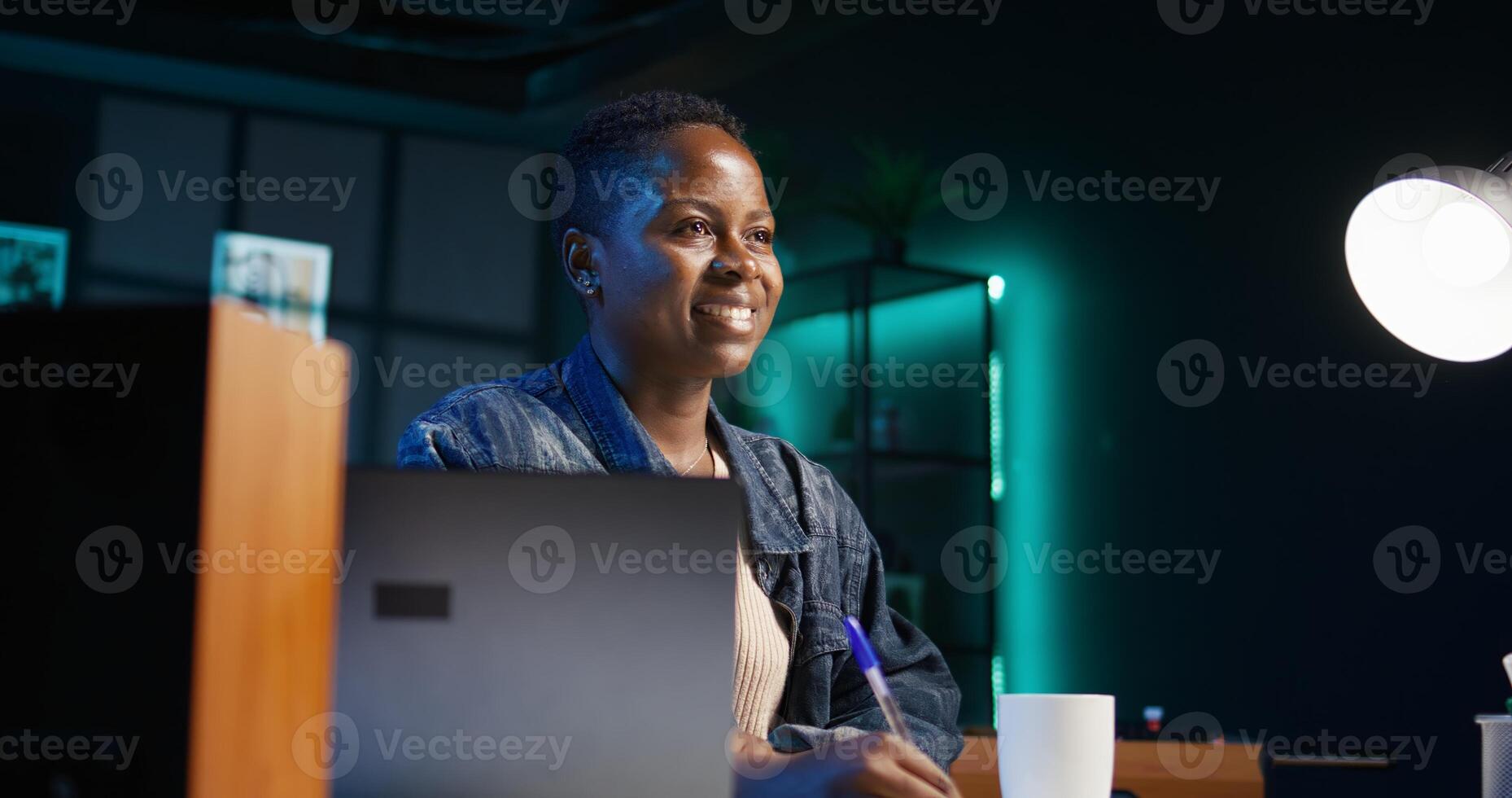 Portrait of smiling woman sitting on chair at home office desk, writing notes on notebook. Cheerful person in living room transcribing information from laptop screen on paper, camera B photo