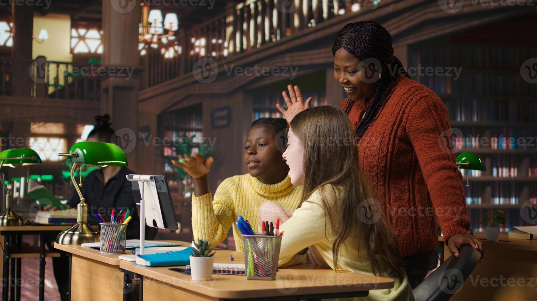 Elementary school pupils and tutor attend a live online course lesson from the library desk, connected via webcam to other classmates in a mentoring session. Virtual webinar discussion. Camera A. photo