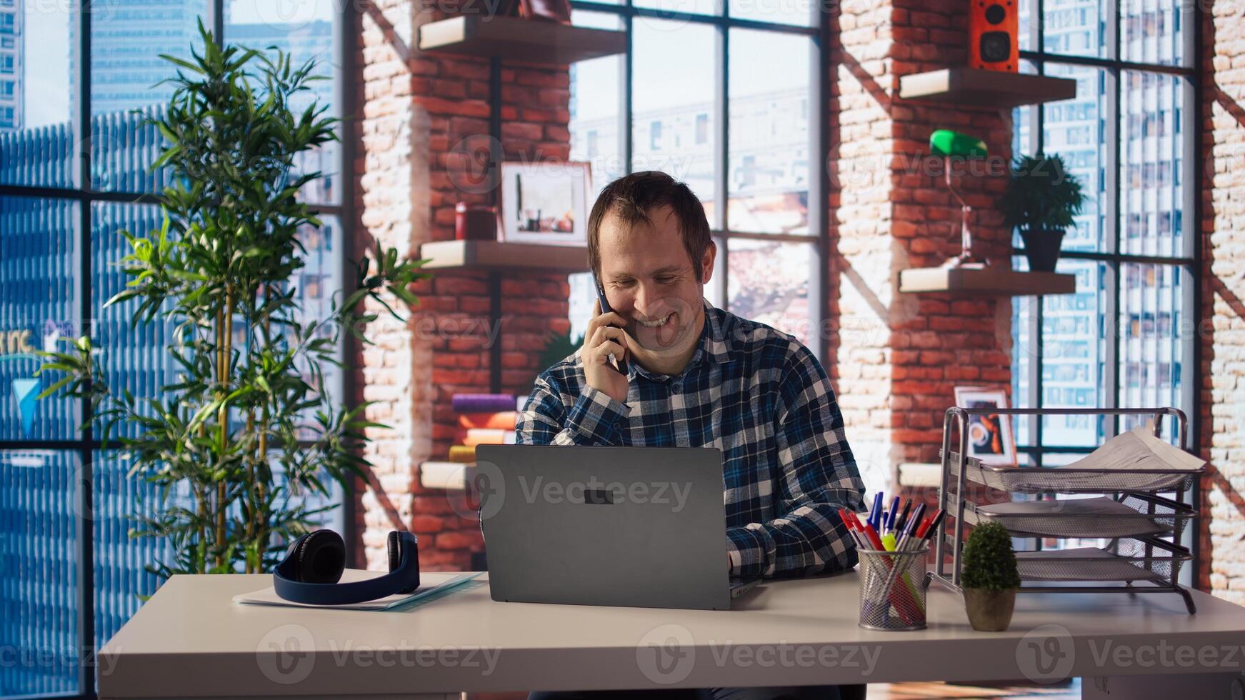 Man seated at home office desk using laptop, answering phone call while checking emails displayed on screen. Worker in telephone call with friend at work while reading messages in inbox, camera A photo