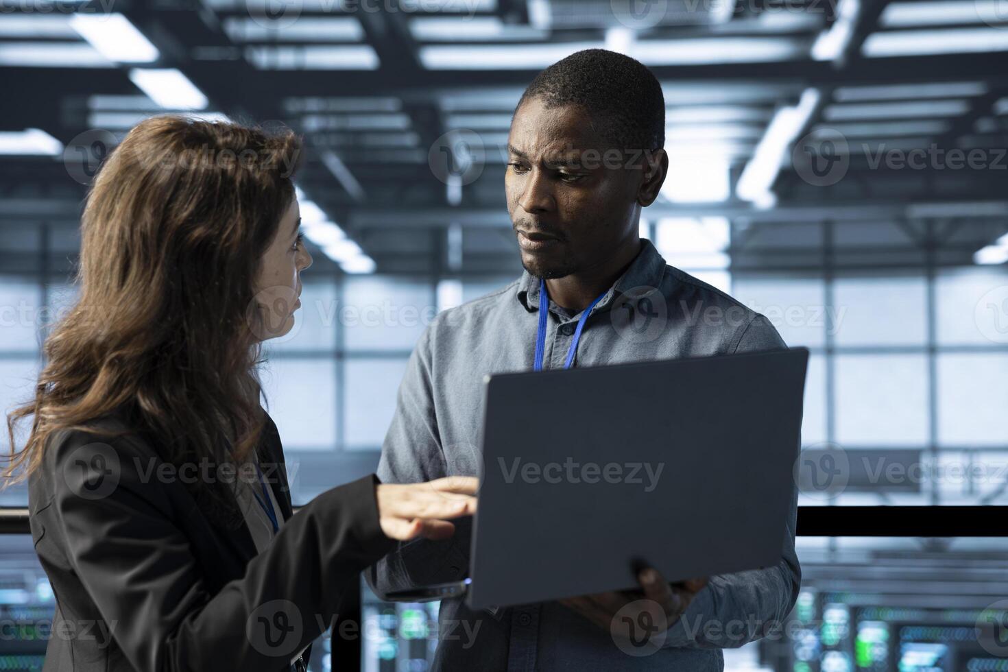 System administrators in data center using laptop, analyzing metrics, optimizing storage volumes. Server farm IT experts using notebook, talking, ensuring seamless operations photo