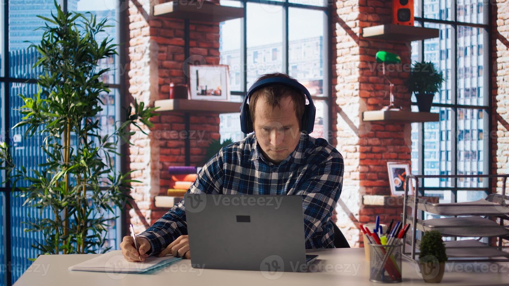Worker listening music, analyzing data on laptop at desk, compiling analytical reports. Employee wearing headphones, using notebook device, writing down information, camera A photo
