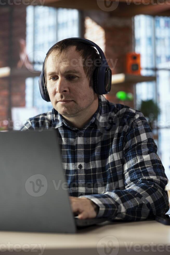 Worker listening music, analyzing data on laptop at desk, compiling analytical reports. Employee wearing headphones, using notebook device, writing down information, finishing project photo