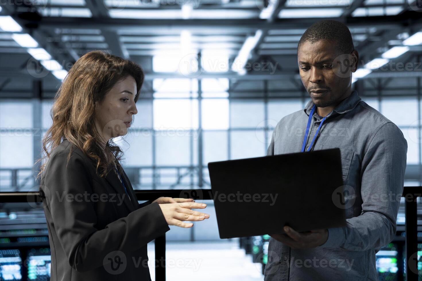Data center programmer and colleague using laptop, conducting security audits, preventing errors. IT staff members cooperating to review firewall rules, logging incident details, talking photo