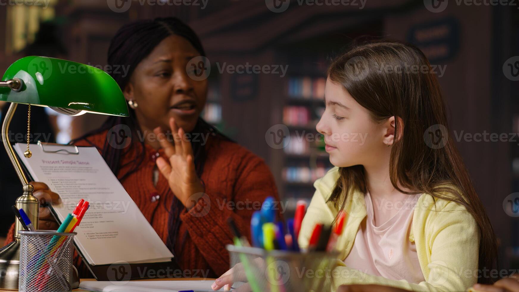 African american professor mentoring diverse schoolgirls in the library, learning together in a library filled with curriculum based materials. Woman tutoring pupils on literature. Camera B. photo