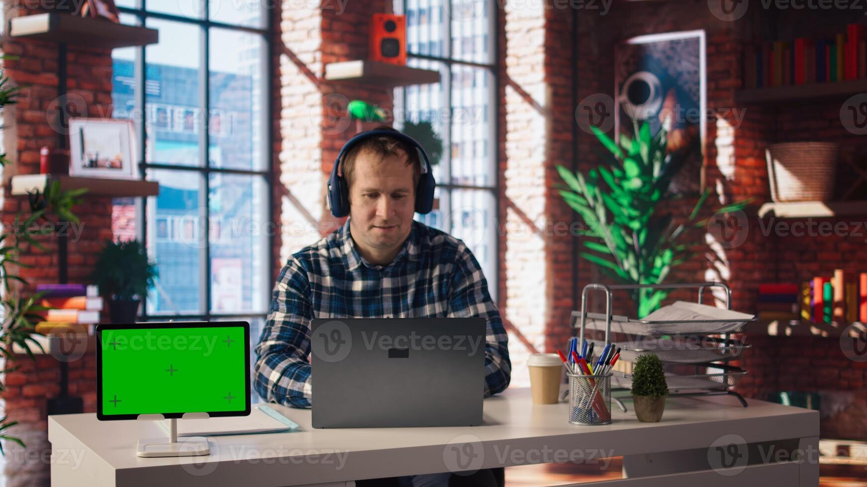 Green screen tablet next to employee completing spreadsheets on laptop and listening music. Chroma key device next to worker in home office wearing headphones editing documents, camera A photo