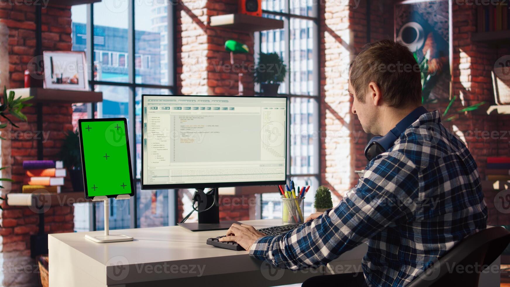 Programmer at desk typing code into computer, developing software programs, consulting green screen tablet. Man in home office fixing errors in application using chroma key device and PC, camera A photo