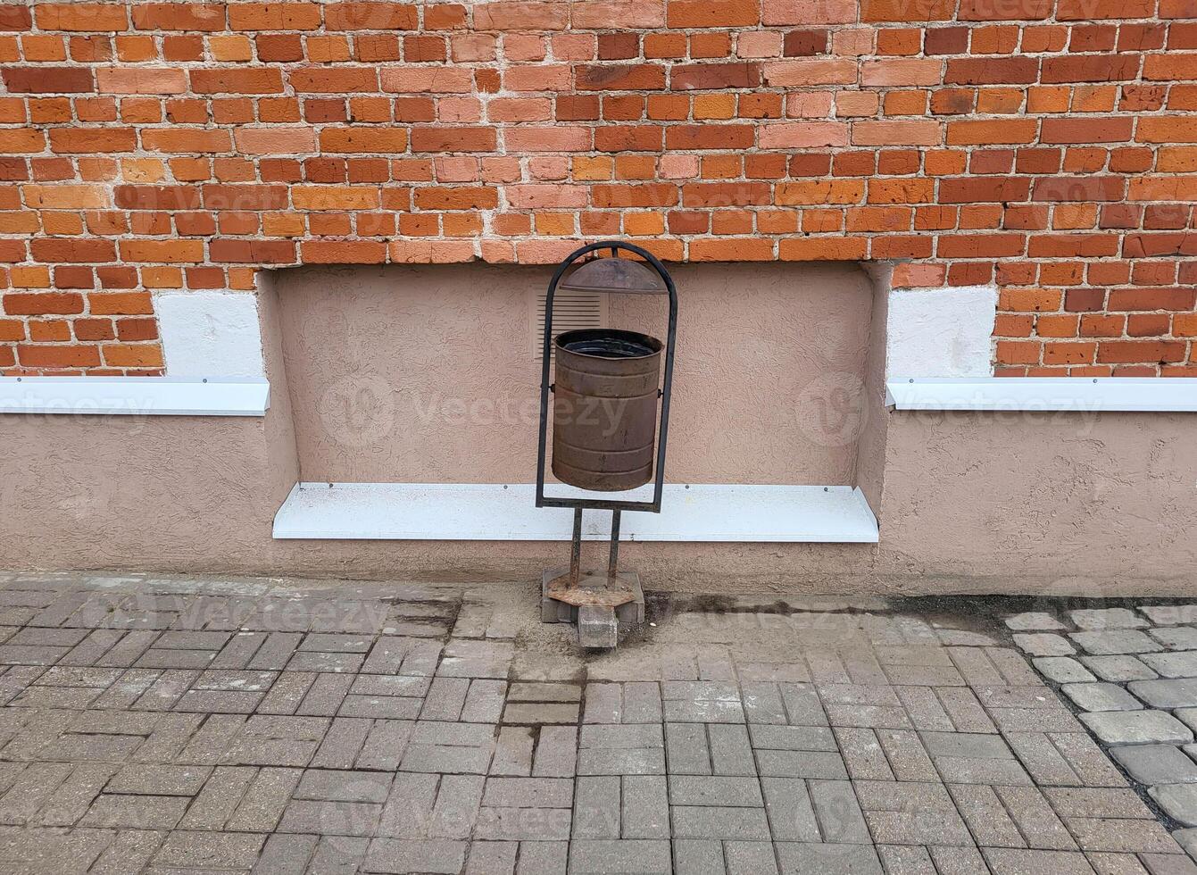 A garbage can on the background of a red brick facade and a pink plastered basement with a ventilation grille photo