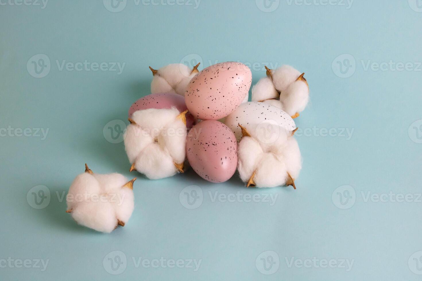 The eggs are the color of Peach Fuzz speckled, lying next to beautiful white cotton flowers on a blue background. The concept of the Easter holiday. photo