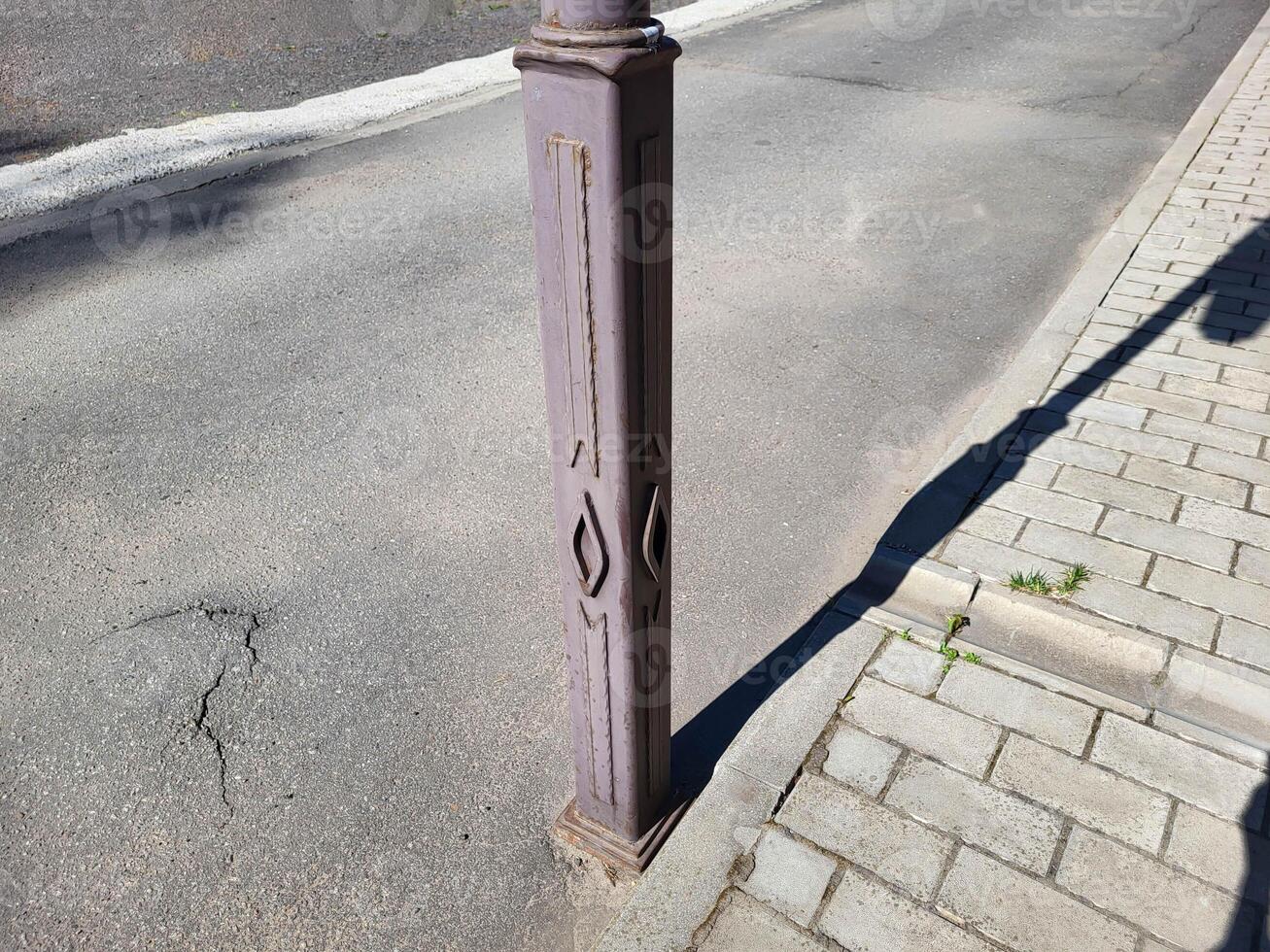On a bright summer day, there is a brown iron pole with forged elements on the street near the sidewalk. photo