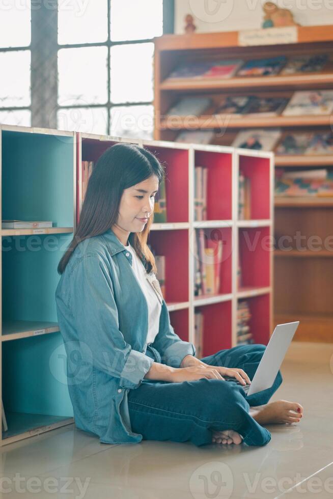 A young woman sits on the floor of a library, working on her laptop, surrounded by colorful bookshelves and natural light. photo