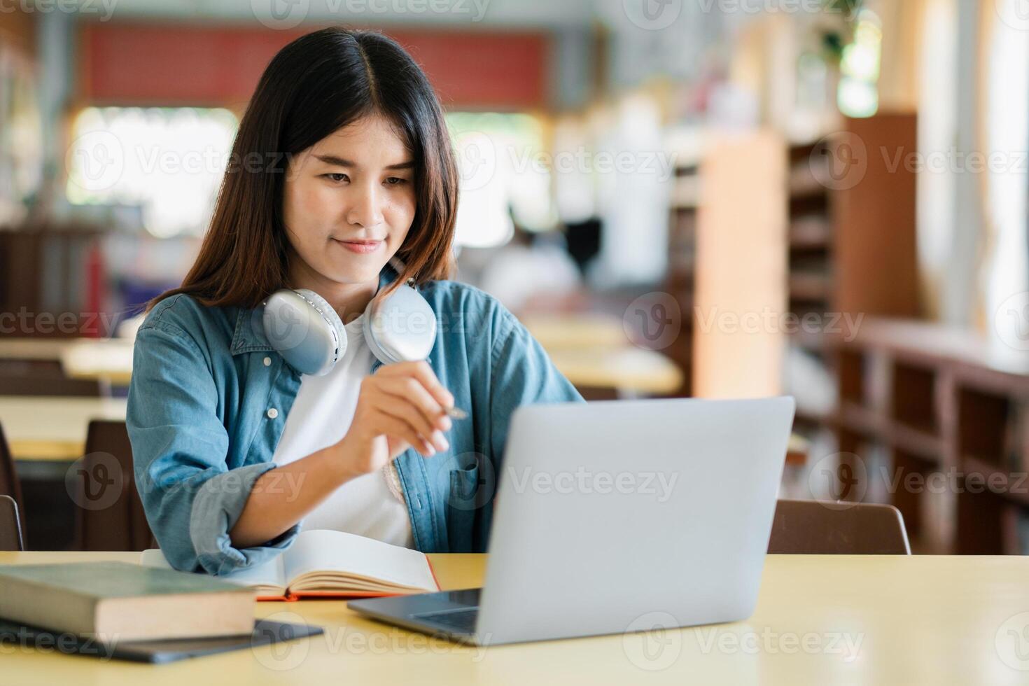 A young woman in a library using a laptop, wearing headphones, surrounded by books, focused on studying and research. photo