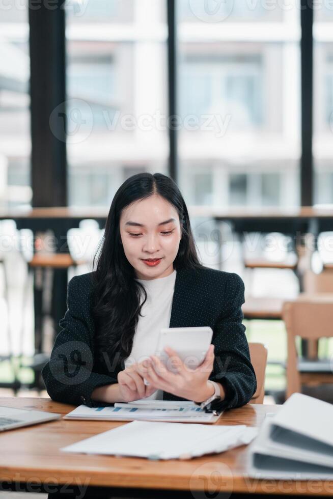 Businesswoman in a modern office using a smartphone, surrounded by documents and a laptop, with large windows in the background. photo
