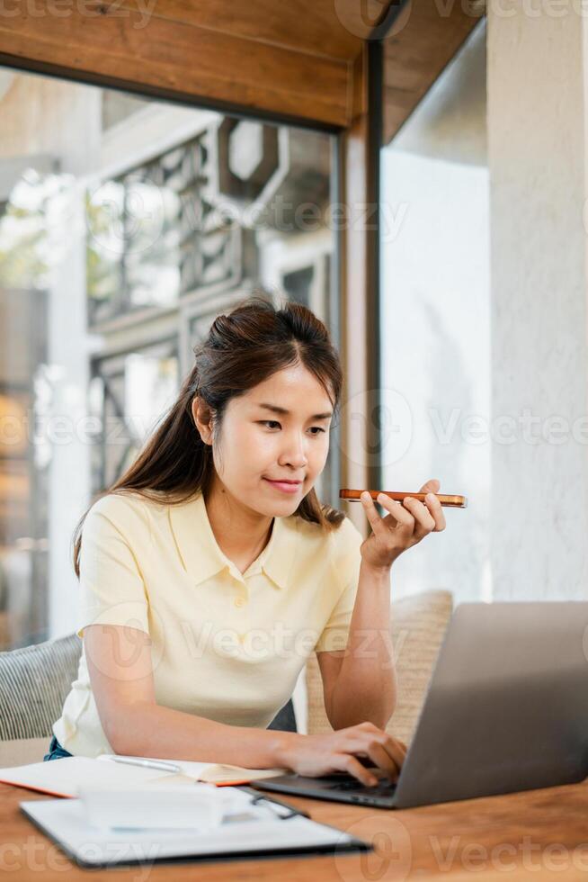 A young woman interacts with her smartphone's voice assistant while working on a laptop in a stylish cafe environment. photo