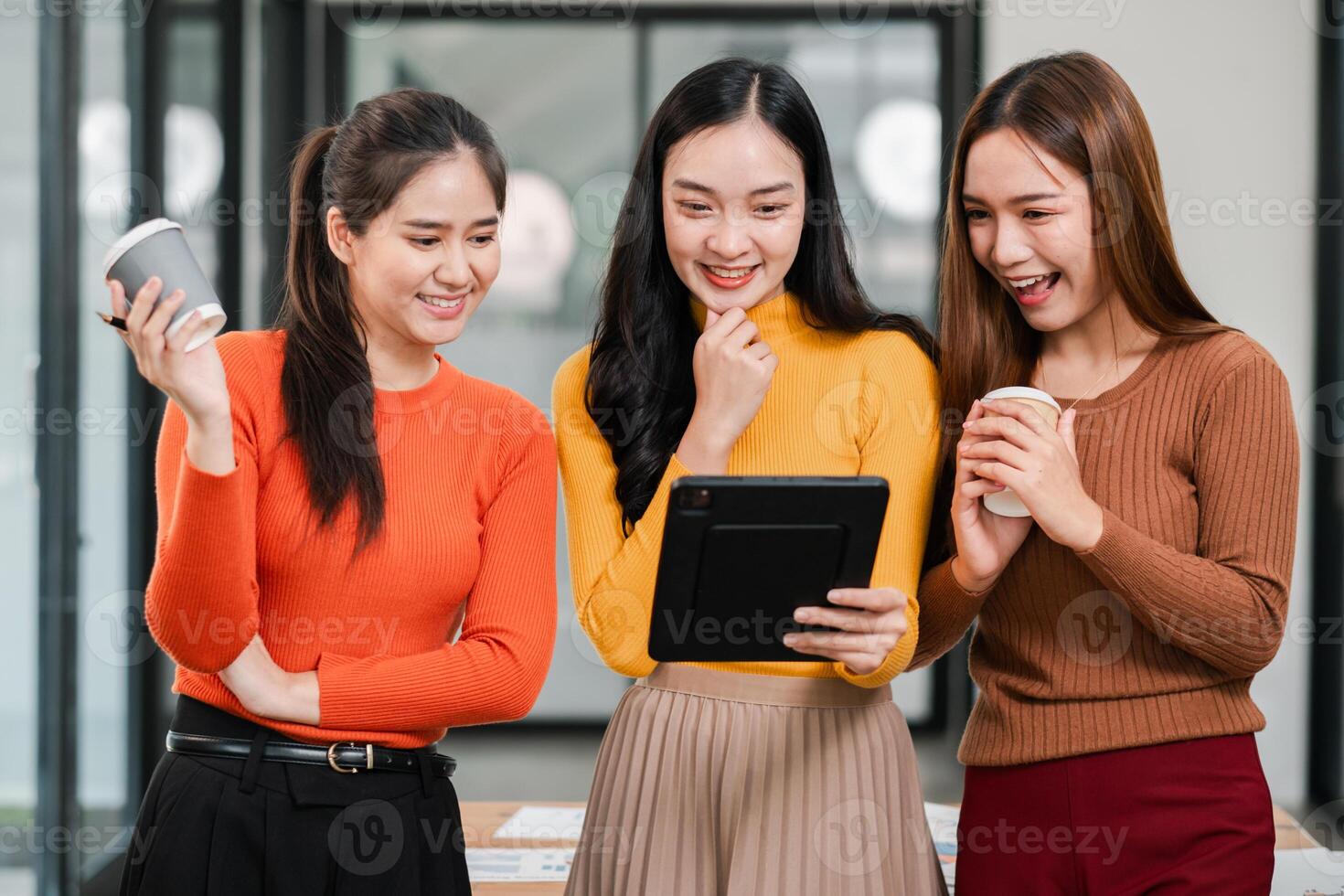 Three young women in a modern office setting, enjoying coffee and engaging with digital content on a tablet. photo