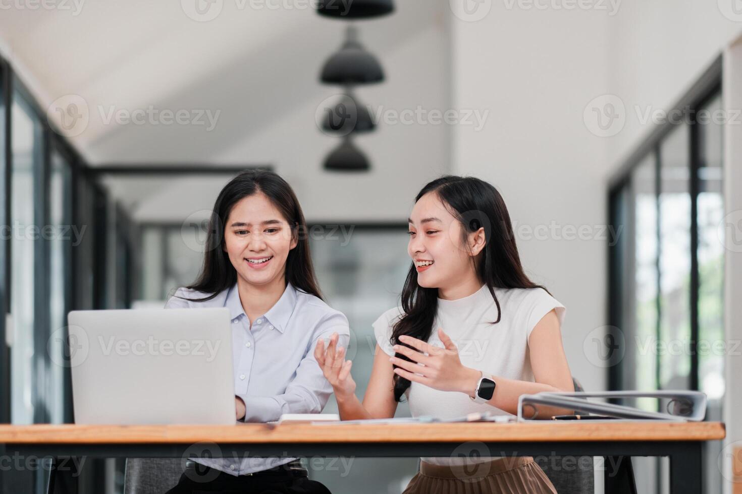 Two young women engaged in a discussion at a modern office table, working on laptops and documents, showcasing teamwork and collaboration. photo