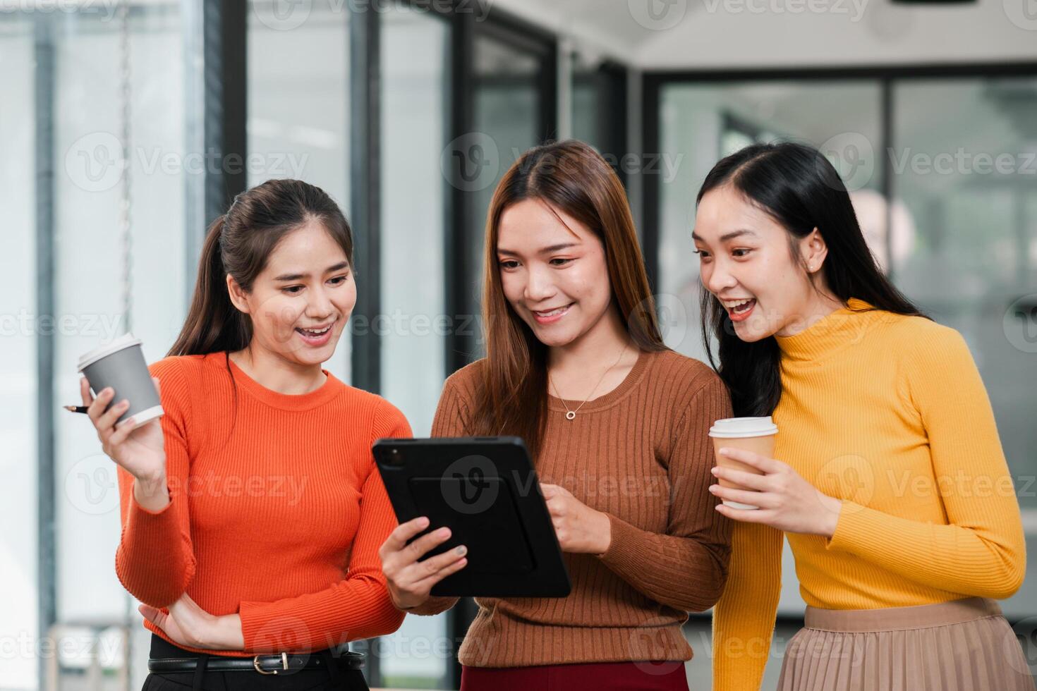 Three women in casual attire share a moment with coffee and a tablet in a bright, modern office environment. photo