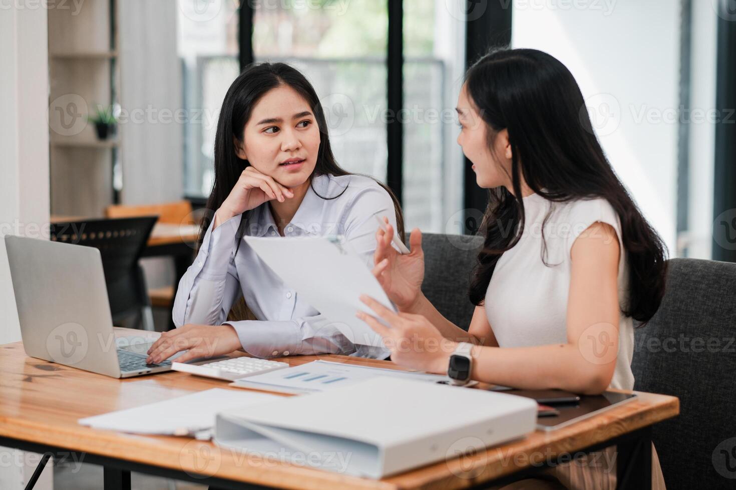 Two women in a modern office setting discussing business strategies with laptops and documents on a wooden table. photo