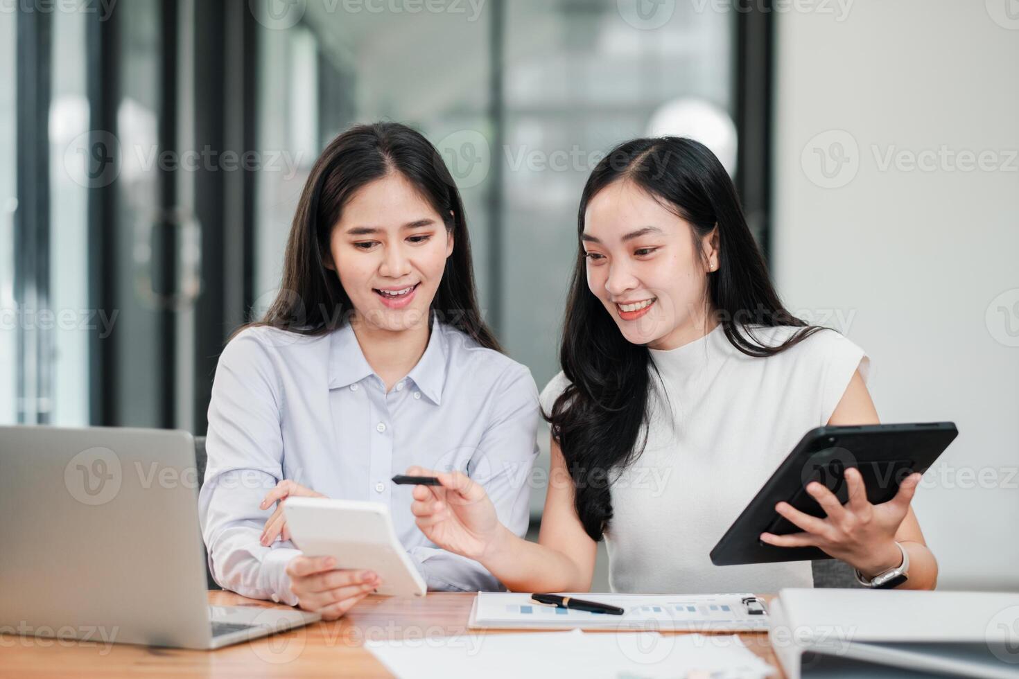 Two women working together in a contemporary office setting, using digital devices and discussing documents. photo
