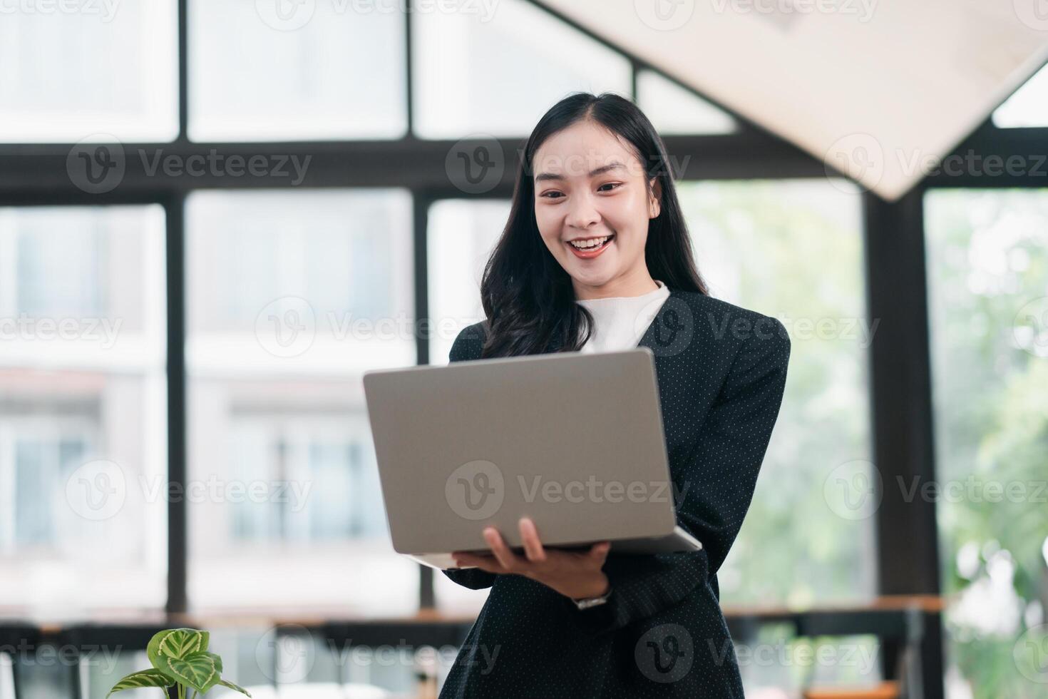 Confident Businesswoman Smiling While Holding Laptop in Modern Office with Large Windows and Natural Light photo