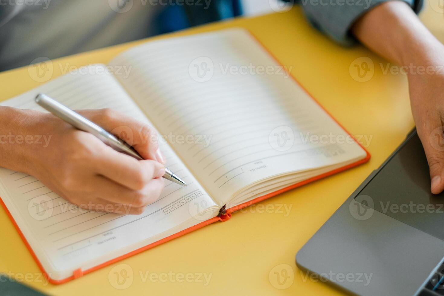 Person Writing in Notebook with Pen Next to Laptop on Yellow Desk in Bright Workspace for Productivity and Creativity photo