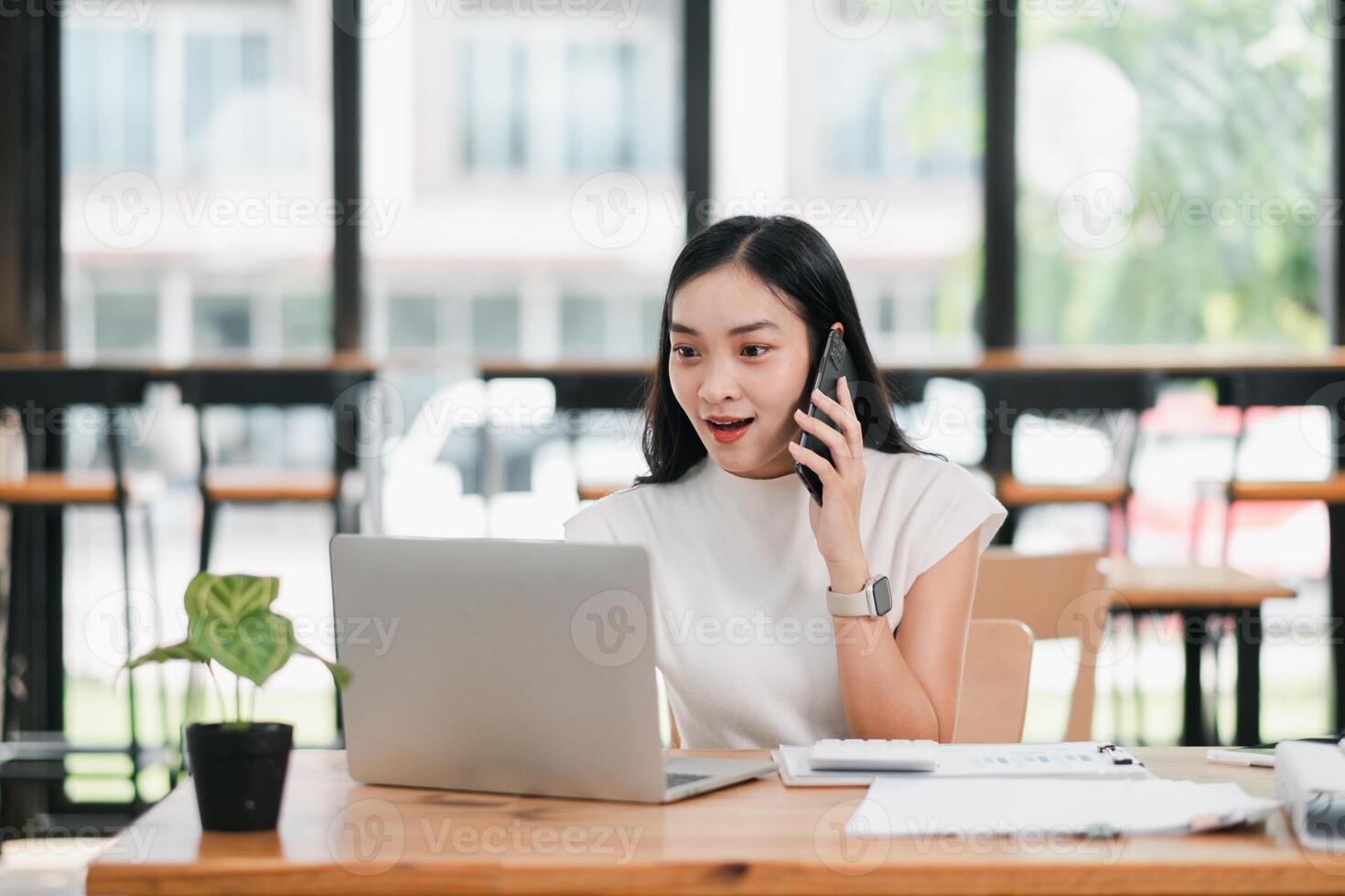 A young woman enthusiastically talks on her smartphone while working on a laptop in a bright, modern office setting. photo