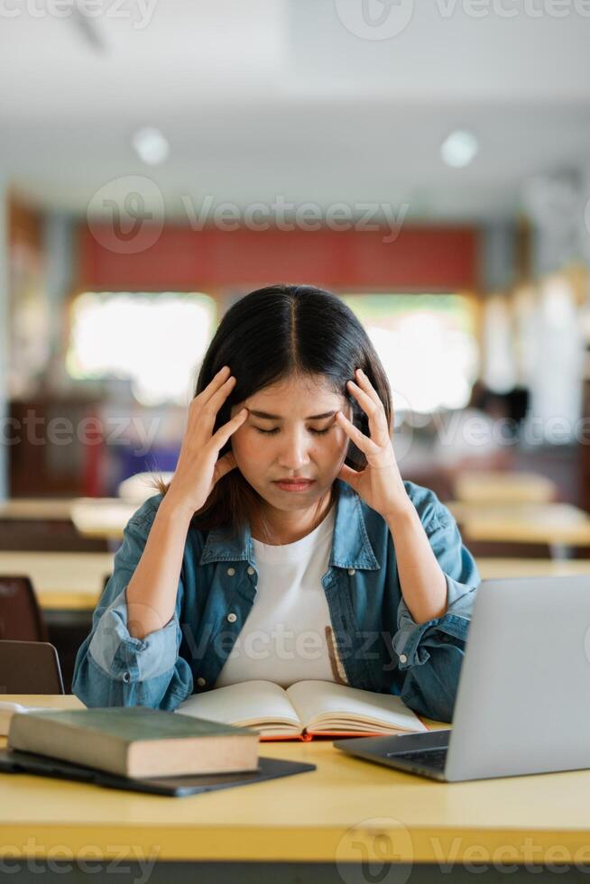 Young Woman Studying in Library with Laptop and Books, Feeling Stressed and Overwhelmed by Academic Workload photo