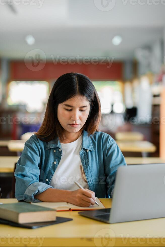 Young woman concentrating on writing notes while using a laptop in a bright study area, showcasing a modern learning environment. photo