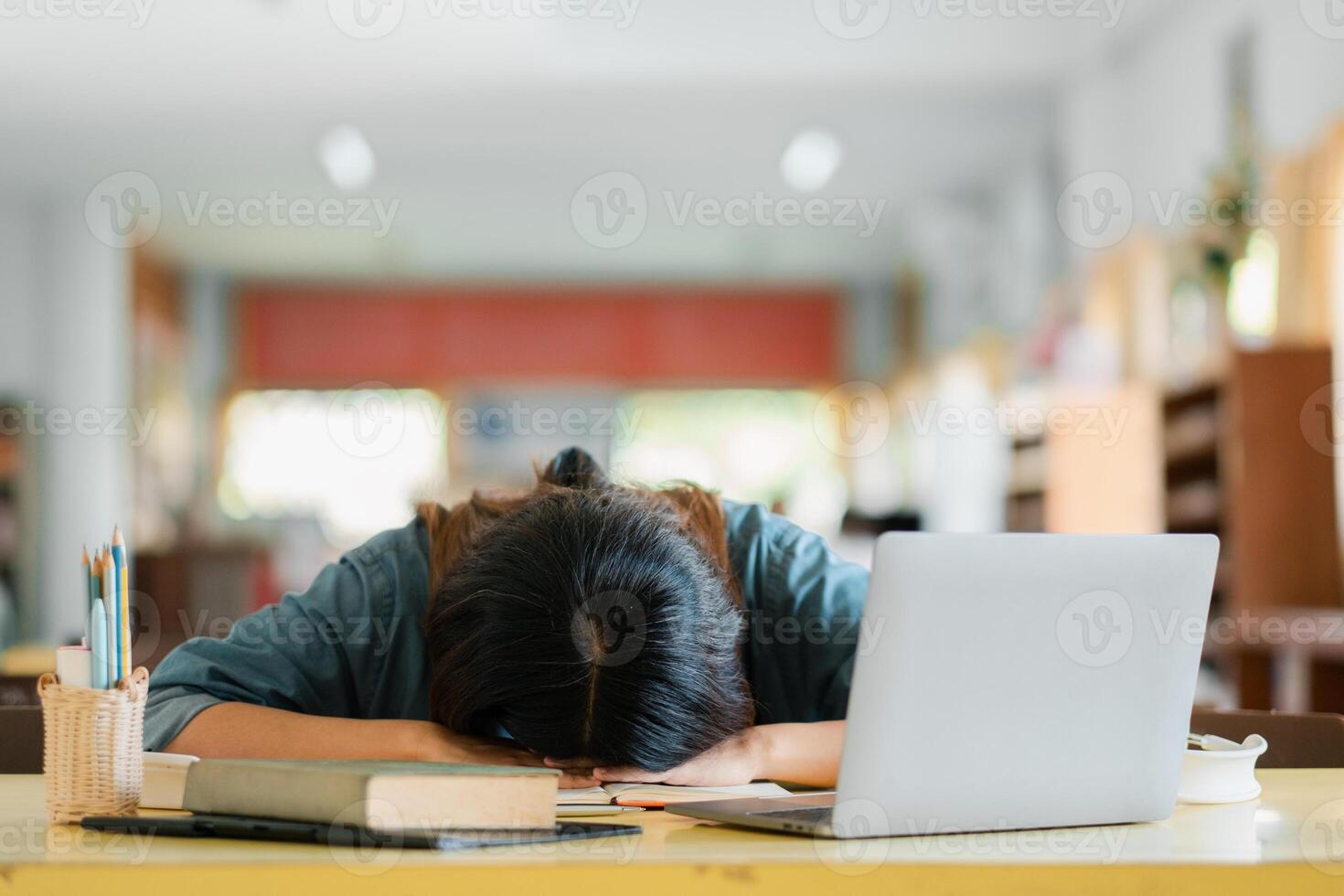 A tired student rests their head on a desk surrounded by a laptop and books in a well-lit classroom environment. photo