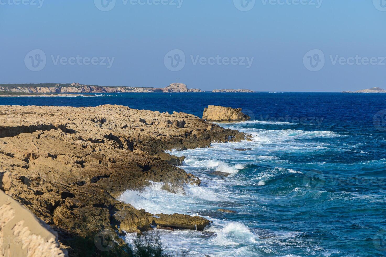 Waves crash against rocky coastal cliffs under a clear blue sky in Cyprus during a sunny day. photo